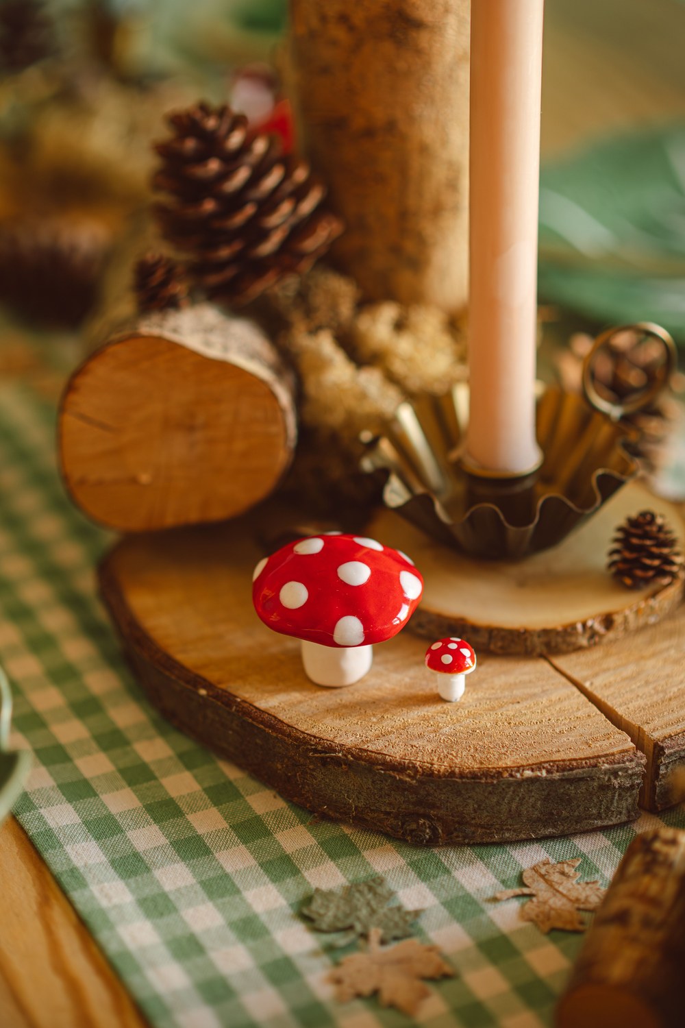 Centre de table en rondins de bois avec champignons rouges en céramique, pommes de pin et bougie sur chemin vichy vert